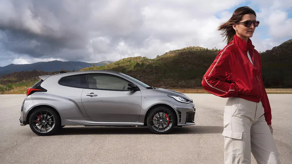 Toyota GR-Yaris en argent, une femme avec une veste rouge, un ciel nuageux et une petite montagne