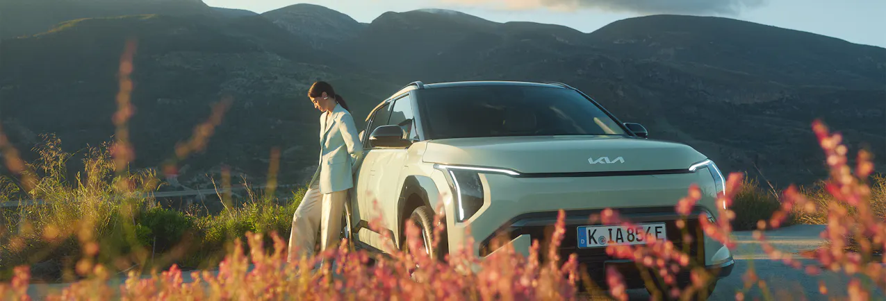 Kia EV3 in Grün, eine Frau lehnt sich ans Auto, Gebirge und Sonnenschein im Hintergrund mit ein paar Wolken