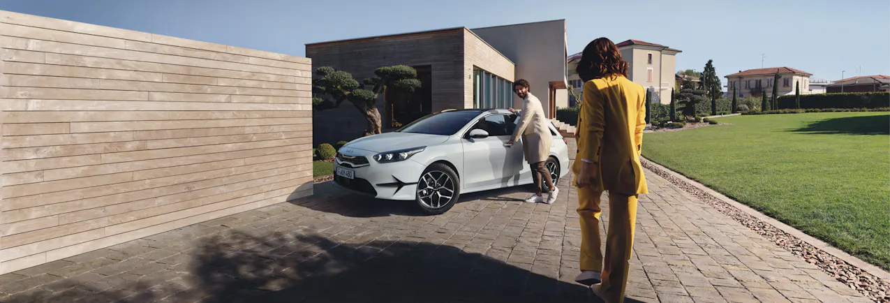 Kia Ceed SW in Weiss, eine Frau in gelber Jacke und ein Mann mit beigen Mantel vor dem Auto, mehrere Häuser und blauen Himmel im Hintergrund