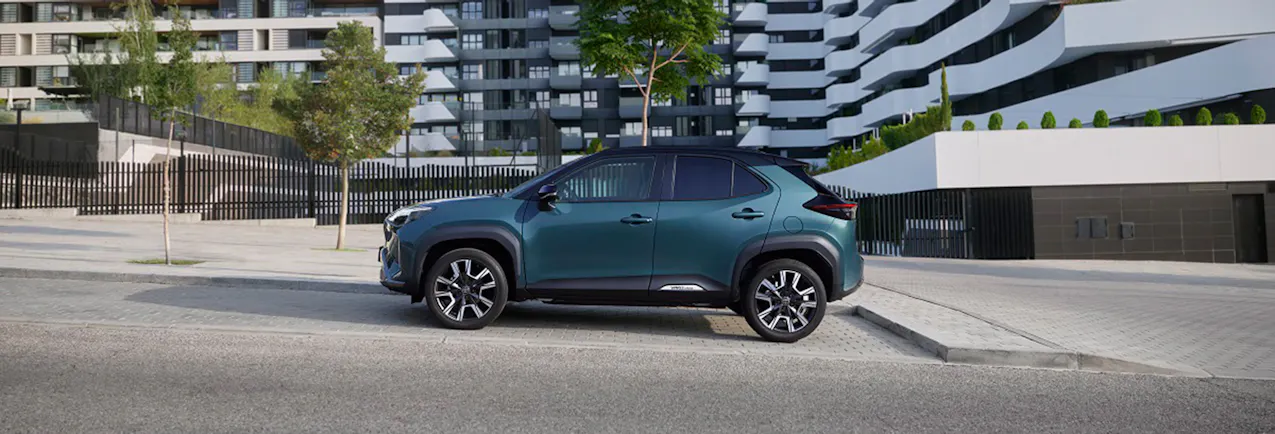 Toyota Yaris Cross in un elegante colore blu, con tre alberi e un moderno complesso residenziale sullo sfondo