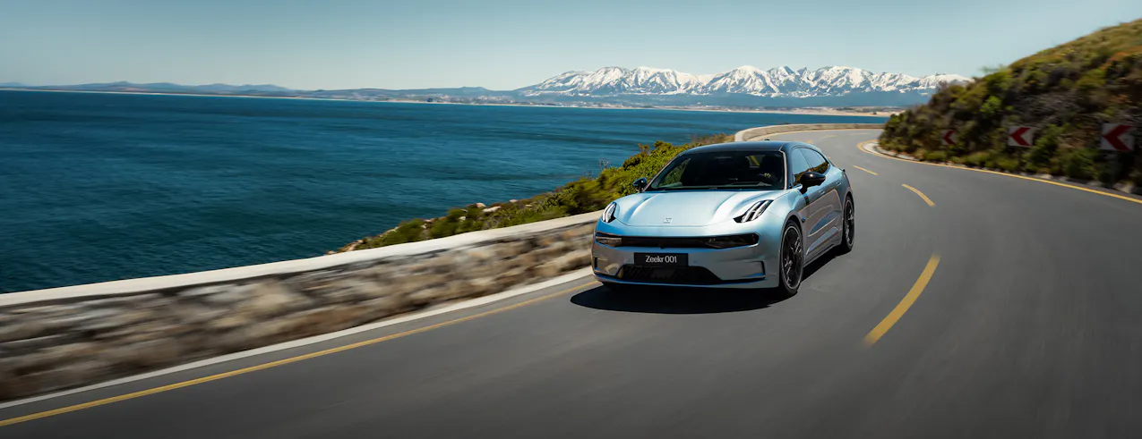 Zeekr 001 en bleu électrique, roulant le long d'un paysage côtier, avec en arrière-plan une chaîne de montagnes sous un ciel clair.