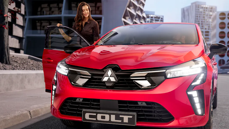 Une femme se tient à côté d'une Mitsubishi Colt rouge, la portière est ouverte, un homme est assis au volant