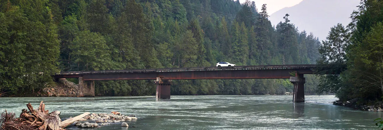 Toyota blanc traversant un pont étroit au-dessus d’une rivière, entouré d’une dense forêt.