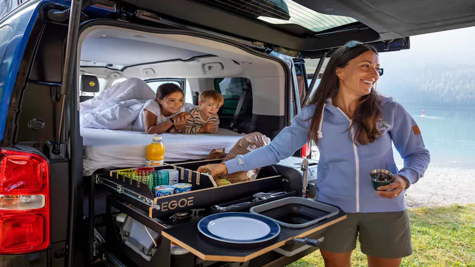 Donna al portellone aperto di un camper Opel con modulo cucina, due bambini sdraiati sul letto, paesaggio idilliaco con lago e foresta