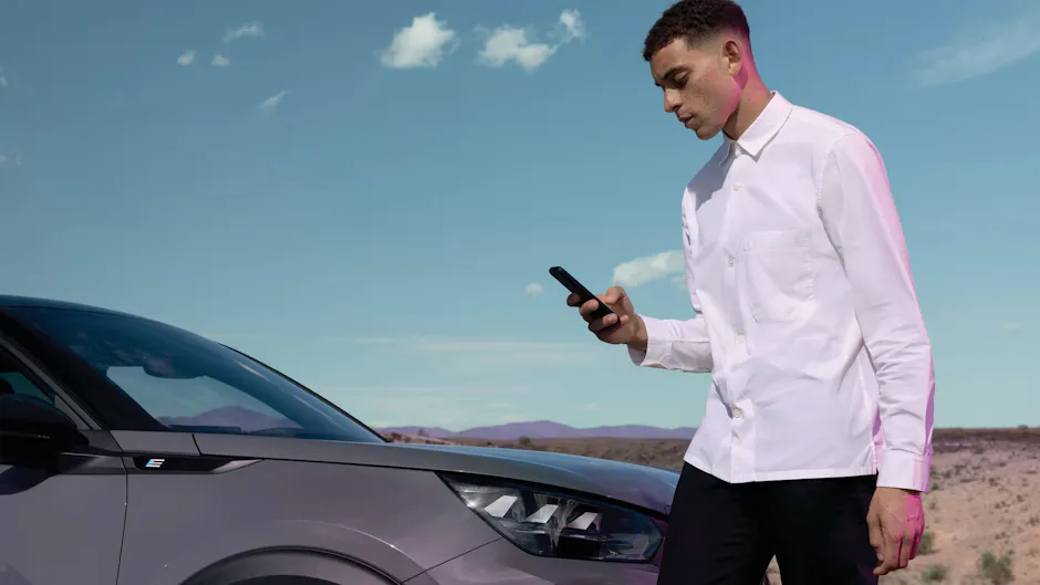 Un homme vêtu d'une chemise blanche, tenant un téléphone portable, devant une Peugeot, avec un paysage désertique en arrière-plan.