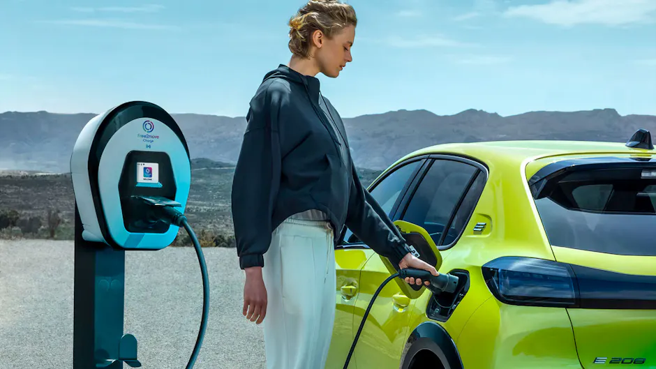 Une femme aux cheveux courts recharge une Peugeot E 208 jaune à une borne de recharge dans une zone désertique.