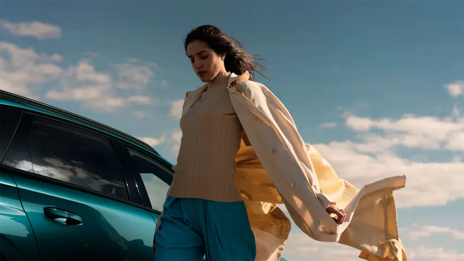 Une femme avec une veste ouverte à côté d’une Peugeot en bleu élégant, sous un beau ciel 