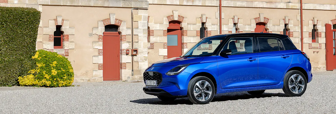 Une Suzuki bleue sur un parking en gravier, avec une belle maison en pierre en arrière-plan.