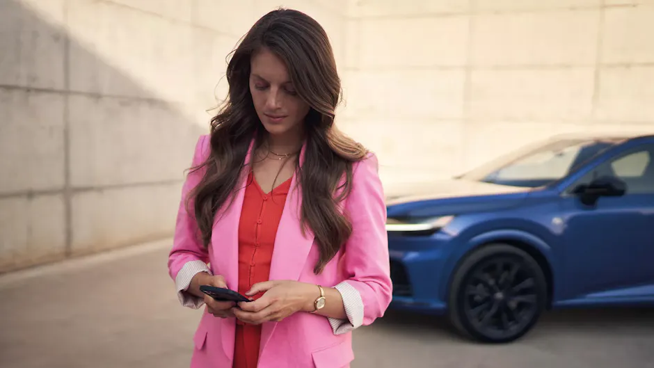 Une femme vêtue d'un blazer rose regarde son téléphone portable, avec une voiture bleue en arrière-plan