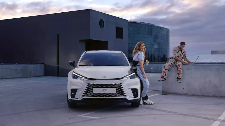 Lexus LBX en blanc, une femme s’appuie contre la voiture, un homme est assis sur un muret et utilise un ordinateur, en arrière-plan un bâtiment futuriste.