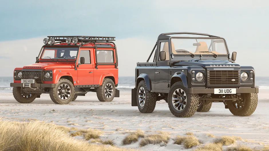 Due Land Rover Defender Classic sono parcheggiati sulla spiaggia, a sinistra un modello rosso con portapacchi e fari supplementari, a destra un modello cabrio nero.