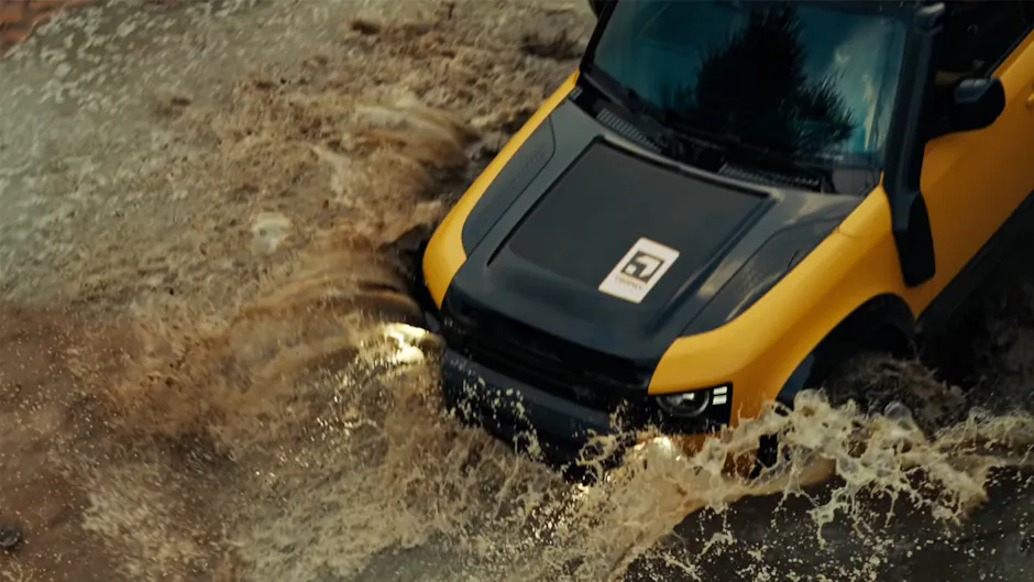 Una Land Rover Defender Trophy Edition color Deep Sandglow Yellow attraversa una pozza d'acqua, ripresa dall'alto