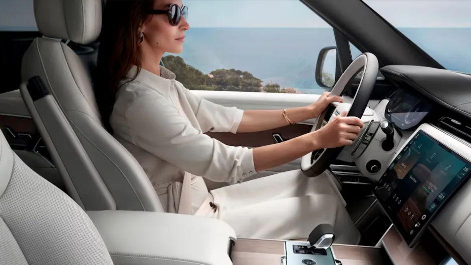 Intérieur blanc d'une Range Rover, une femme portant des lunettes de soleil est assise au volant, la mer en arrière-plan.