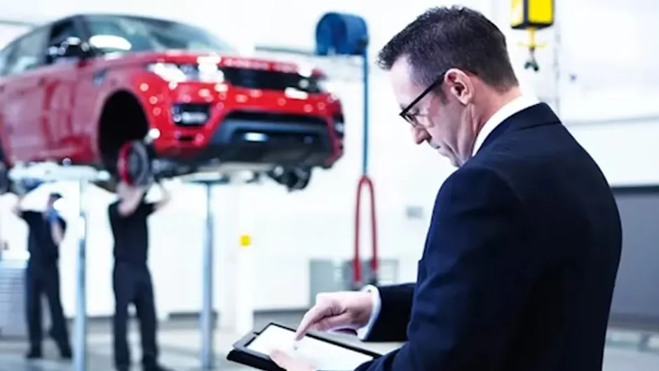 Eine Person im Anzug bedient ein Tablet, ein roter Range Rover ist auf der Hebebühne in der Garage, zwei Personen arbeiten unter dem Auto