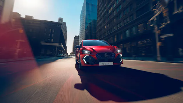 Roter Renault Clio fährt frontal durch urbane Innenstadt mit moderner Architektur, dynamische Fahraufnahme.
