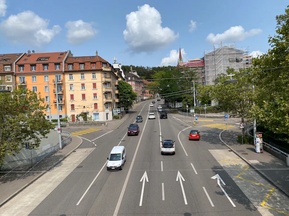 Vista dall'alto di un'ampia strada cittadina a più corsie, con automobili nel traffico, edifici residenziali, alberi e cielo blu con nuvole in un contesto urbano.