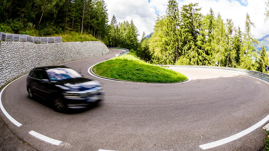 Un'auto nera percorre una curva su una strada di montagna; sullo sfondo si vedono alberi verdi