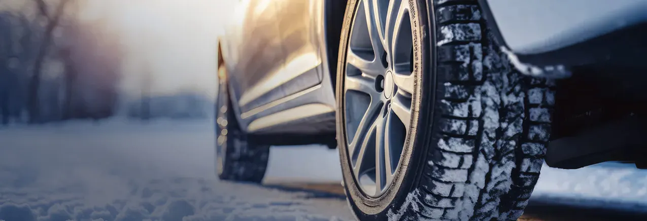 Un'auto è parcheggiata sulla neve; l'attenzione è concentrata sugli pneumatici e sul battistrada invernale, mentre sullo sfondo sorge il sole